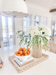 Woven basket tray styled with white hydrangeas in a rattan vase, a bowl of oranges used as bowl filler, and stacked coffee table books. A perfect example of capsule home decor for a bright and airy kitchen.