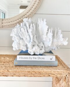 Coastal-inspired decor featuring a white coral sculpture stacked on coffee table books, styled on a woven console table. A perfect blend of natural textures for a modern coastal home.