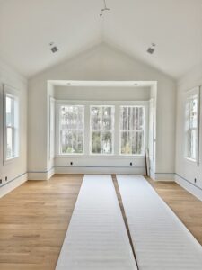 Bright bedroom under construction featuring vaulted ceilings, large windows overlooking wooded view, and light oak hardwood floors.
