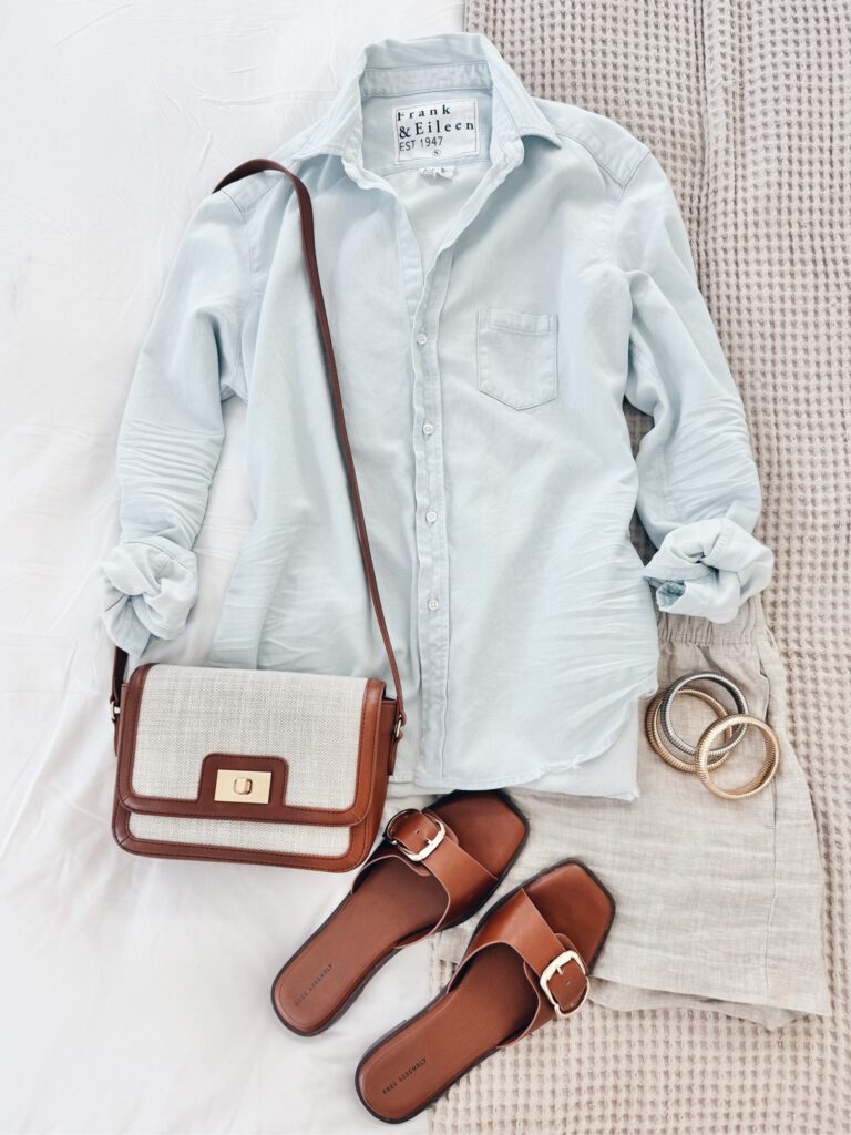 Relaxed summer outfit with a pale blue Frank & Eileen button-up shirt, beige linen shorts, tan slide sandals, a neutral crossbody bag with gold hardware, and gold bangles.