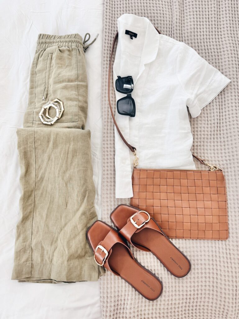 Casual summer outfit featuring a white short-sleeve linen shirt, olive green linen joggers, tortoiseshell bangles, square black sunglasses, a tan woven crossbody bag, and tan slide sandals with gold buckles.